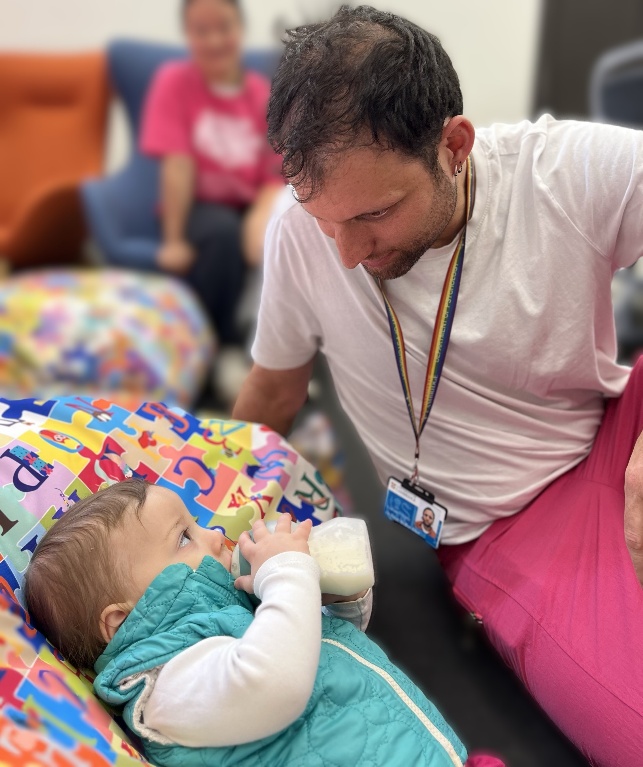 A volunteer watches on as a baby leans against a brightly-coloured cushion and drinks a bottle, in the feeding area.